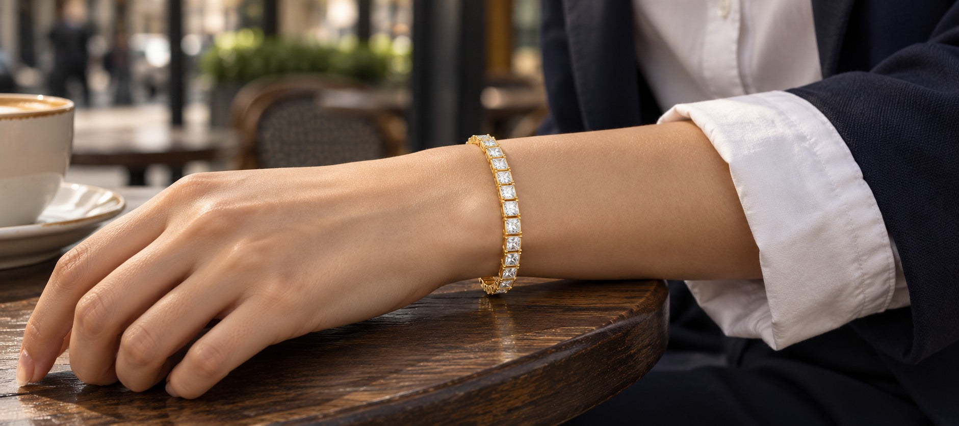 Gold square-stone tennis bracelet worn with a white shirt and navy jacket at a cafe